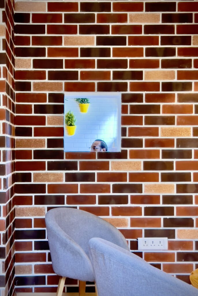 Photography of a brick wall with a small window opening, indoor plants, a partial human presence, and a grey chair in the foreground.